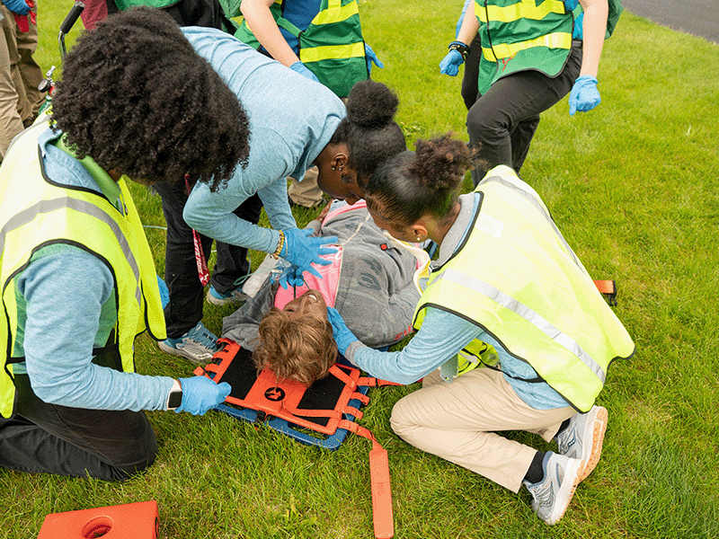 Students practicing emergency medical response on a simulated injured person on grass during training